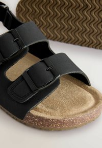 Sort sandal med to brede remme med spænder, polstret suede fodseng og struktureret gummisål med et bølget mønster.