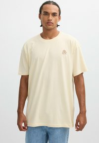 Camiseta de algodón beige con un corte relajado, mangas cortas y un pequeño bordado de rosa en el lado izquierdo del pecho. Lleva puesta unos vaqueros de color azul claro.