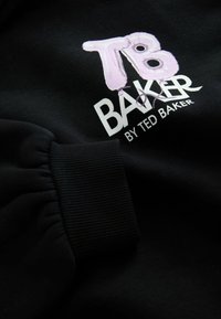 Čierna mikina s nariaseným rukávom a bielym a ružovým textovým logom "TB BAKER" od Ted Bakera, s hladkým povrchom látky.