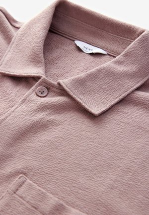 Polo de couleur rose pâle avec un tissu texturé, doté d'un col, d'une fermeture à boutons et d'un détail de poche discret. L'étiquette indique "NEXT".