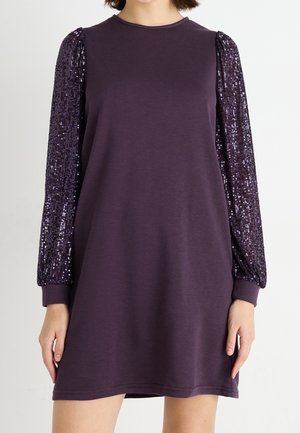 Woman wearing a purple long-sleeve dress with sequined sleeves and a plain body, standing against a neutral background.