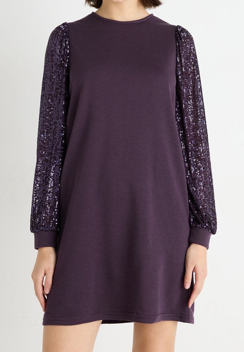 Femme portant une robe violette à manches longues avec des manches à sequins et un corps uni, debout devant un fond neutre.