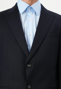 Marinefarbener Textilblazer mit zwei Knöpfen und einer einzigen Brusttasche. Darunter ein hellblaues Hemd mit Kragen und Perlmuttknöpfen.