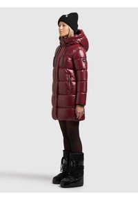 Abrigo acolchado burdeos con capucha, acabado brillante, detalles en negro, dos bolsillos delanteros y cierre de cremallera. Combinado con botas de invierno negras.