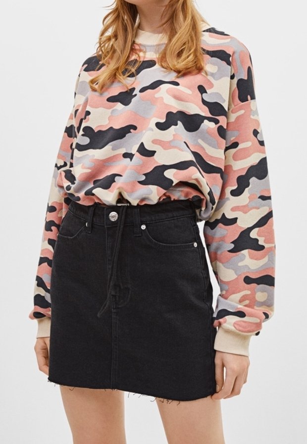 Femme portant un sweat camouflage rose pastel, noir et beige, rentré dans une jupe en jean noire avec poches avant et ourlet brut.