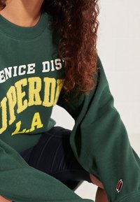 Woman wearing a green oversized sweatshirt with yellow and white "Venice District Superdry L.A." text and dark pants, sitting against a white background.
