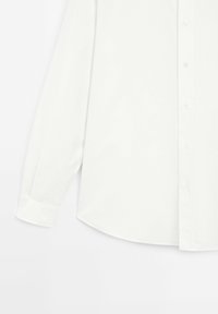 Chemise blanche à manches longues avec col standard et poignets boutonnés, étalée à plat sur un fond blanc.