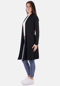 Cardigan lungo nero con apertura frontale, realizzato in un materiale morbido. Presenta maniche lunghe e due tasche laterali. Indossato sopra una maglietta bianca.