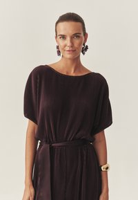 Szilva színű, plisszírozott ruha széles, bardot nyakkivágással, laza ujjakon és passzoló övvel. Arany karpereccel és virág fülbevalókkal díszített. Sima tapintású.