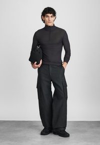 Top preto de manga longa com fecho, combinado com calças cargo pretas de corte largo e calçado preto. Modelo segura uma pequena bolsa preta.