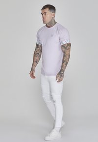 Gaiši violets pielāgots T-krekls ar KSI logotipu uz piedurknēm, saskaņots ar nolietotām baltām šaurajām džinsām un baltām sporta apaviem. Redzamas tetovējumi uz rokām.