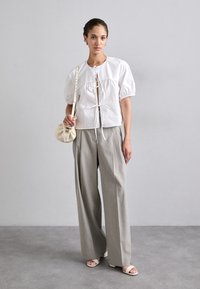 Blouse blanche à manches courtes avec détail de nœud à l'avant, associée à un pantalon large taille haute gris clair et des sandales légères. Sac crème à la main.