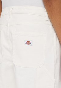 Pantalones de trabajo de mezclilla blanca con un bolsillo trasero y una etiqueta de logo colorida de Dickies. Tejido resistente con una textura suave y diseño liso.