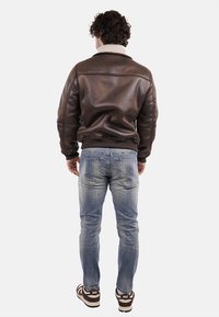 Giacca bomber in pelle marrone con collo in shearling, design aderente e polsini a costine. Indossata con jeans chiari strappati e sneakers.