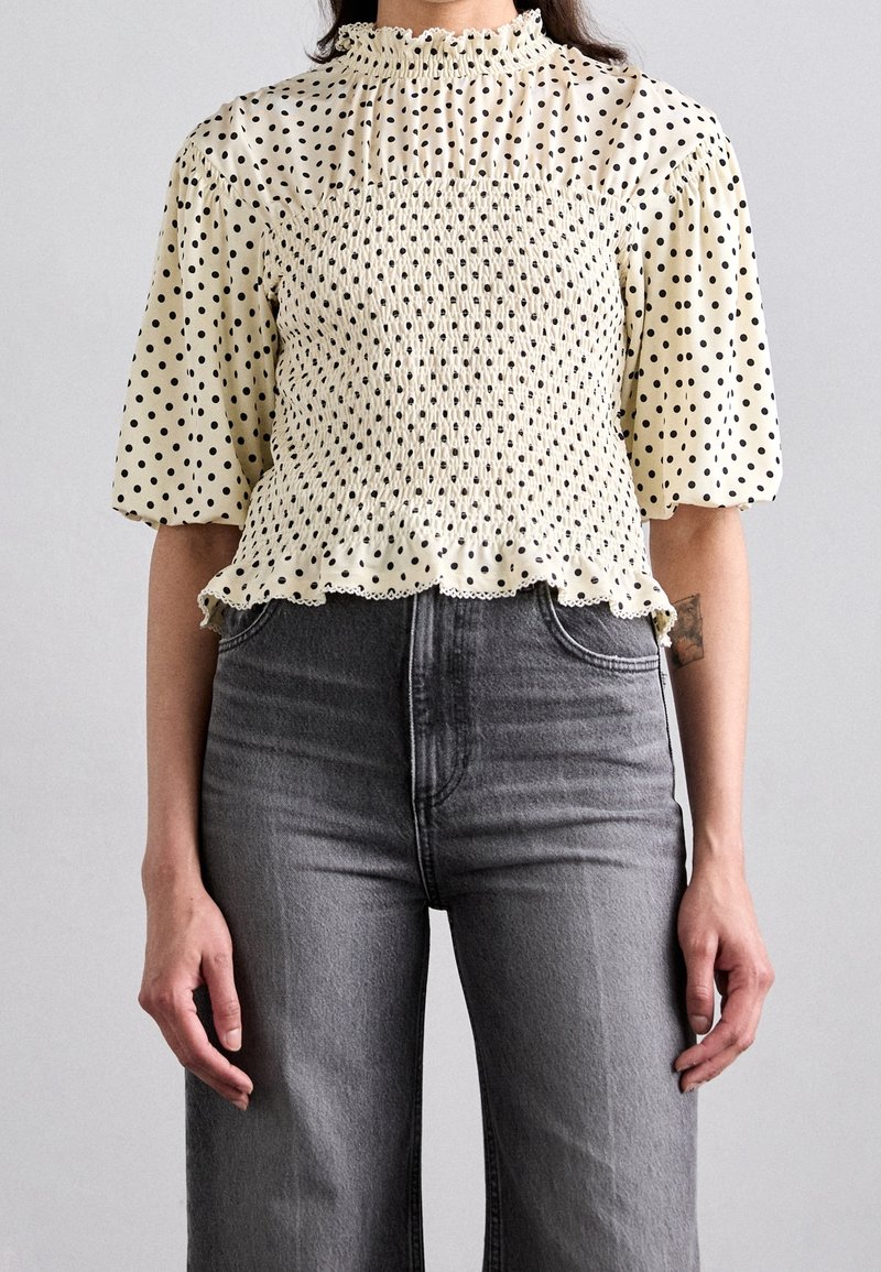 Femme portant un chemisier smocké couleur crème à pois noirs avec des manches mi-longues bouffantes, associé à un jean gris taille haute.