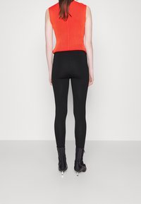 Leggings noirs associés à un top sans manches côtelé orange, présentant une silhouette ajustée et une texture lisse. Le mannequin porte des bottines à talons noires.