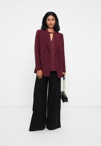 Blazer duplo em bordeaux com botões pretos, combinado com uma blusa a condizer e calças largas pretas; segurando uma pequena mala preta.