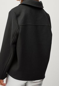 Blouson noir avec une coupe décontractée, présentant un grand col, une texture lisse et un détail de couture dans le dos.