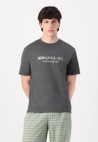 Szürke, rövid ujjú póló fehér grafikai mintákkal, amelyek között szerepel egy vonalkód, teknős és japán szöveg. Az anyag simának és könnyűnek tűnik.