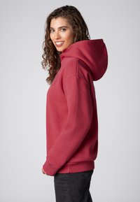 Pullover hoodie van dieprood katoen, met een grote capuchon, geribde boorden en een geborduurd logo op de borst. Het model draagt zwarte jeans.