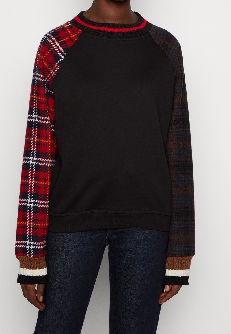 Personne portant un sweat-shirt noir avec une manche gauche à carreaux rouges, une manche droite à carreaux sombres et des poignets rayés, associé à un jean bleu foncé.