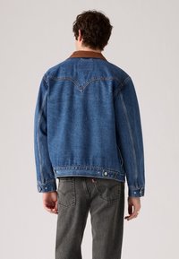 Hombre que lleva una chaqueta de mezclilla azul con cuello marrón y jeans negros desgastados, de espaldas contra un fondo simple.