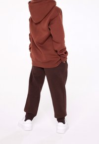 Felpa con cappuccio marrone dal taglio rilassato, abbinata a pantaloni della tuta marrone scuro. Sneakers bianche accentuano l'outfit. Texture morbida in tutto il capo.