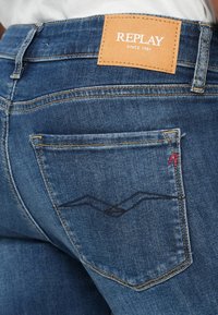 Jean en denim bleu avec une étiquette en cuir beige arborant "REPLAY" et "SINCE 1981". La poche arrière présente un design brodé en bleu foncé.