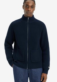 Cárdigan de punto acanalado azul marino con cremallera, que presenta un alto collar, dos bolsillos frontales y un acabado texturizado. Diseño simple y moderno.