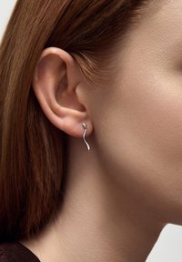 Srebrne kolczyki o falistym wzorze, z gładkim, wypolerowanym wykończeniem. Przymocowane do płatka ucha, mają smukły, elegancki kształt.