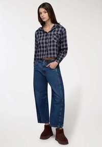 Giovane donna in piedi con una mano in tasca, indossa una camicia a quadri blu scura cropped, jeans a gamba larga blu e stivaletti marroni su sfondo bianco.