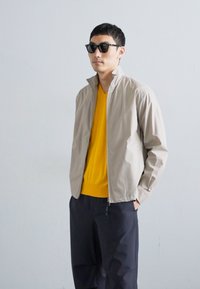 Lys beige zip-up jakke med ståkrage, brukt over en lys gul genser og mørke bukser. Glatt stoff og minimalistisk design.