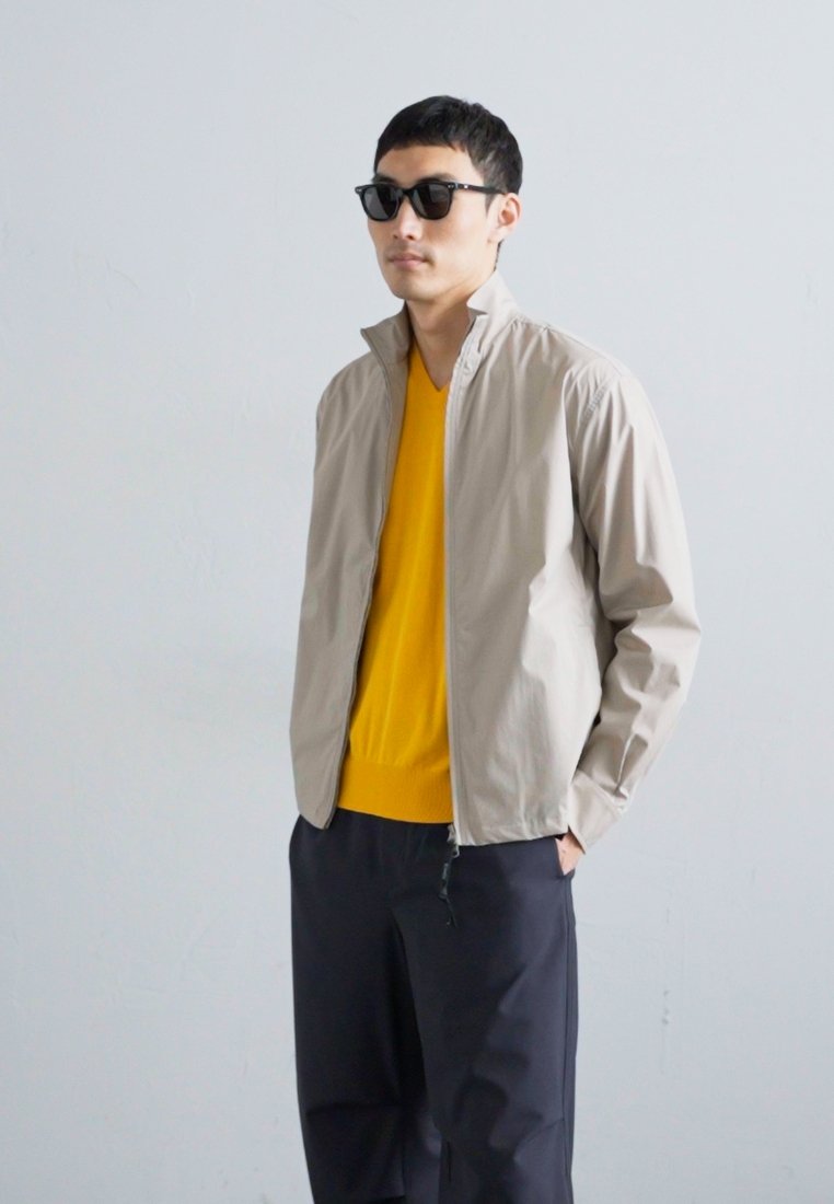 Light beige zip-up jacket with a stand collar, worn over a bright yellow sweater and dark trousers. Smooth fabric and minimalistic design.