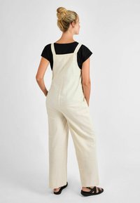 Salopette in lino beige leggero con gambe larghe, spalline regolabili e un taglio rilassato, abbinata a una maglietta nera a maniche corte e sandali.