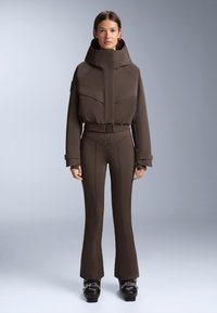 Ruskea hiihtotakki, jossa korkea kaulus ja etutaskut, yhdistetty istuviin ruskeisiin housuihin, joissa on saumoja. Asun täydentävät mustat hiihtomonot.