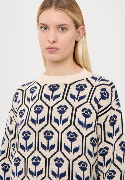 Pull en tricot beige avec un motif floral hexagonal en bleu foncé et marine. Encolure ronde avec bords côtelés, texture de poids moyen.