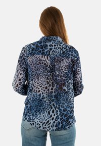 Persoon met lang bruin haar draagt een blouse met een blauw en zwart luipaardprint en lichte blauwe jeans, van achteren weergegeven tegen een witte achtergrond.