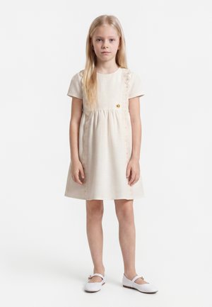 Jeune fille aux longs cheveux blonds portant une robe crème à manches courtes et des chaussures Mary Jane blanches, debout devant un fond uni.