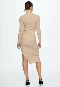 Robe en maille côtelée beige avec un col haut, des manches longues et une fente sur le côté, assortie à des talons noirs, mettant en valeur une silhouette ajustée.