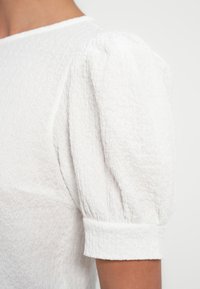 Blouse blanche texturée avec manches courtes bouffantes et revers roulés. Le tissu présente une finition subtilement froissée, ajoutant de la profondeur au design.