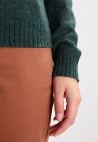 Grüner Wollpullover mit gerippten Bündchen, kombiniert mit einem braunen Stoffrock. Fokus auf die weiche Textur und die kontrastierenden Farben.