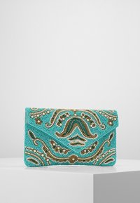 Clutch di perline turchesi con dettagli floreali e a spirale intricati in oro e bianco, appoggiata su un piedistallo bianco contro uno sfondo grigio.