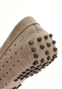 Beige suède schoen met een gestructureerde afwerking, voorzien van kleine bruine rubberen nopjes op de zool voor grip, en ventilatiegaten langs de zijkanten.