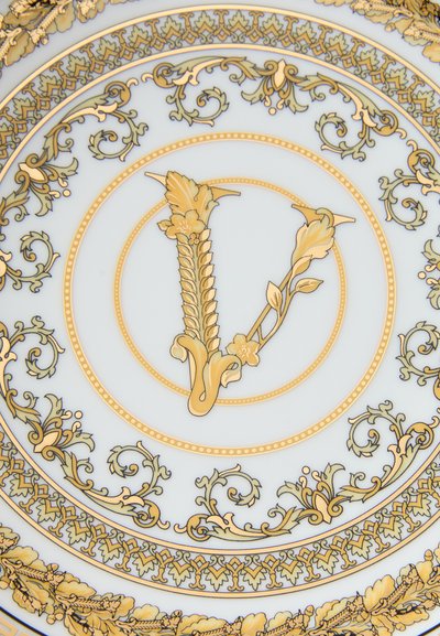 Porcelāna šķīvis ar sarežģītiem zelta ziedu rakstiem un centrālu zelta "V" dizainu uz balta fona, ieskauts dekoratīvā vērpšanas rotā.