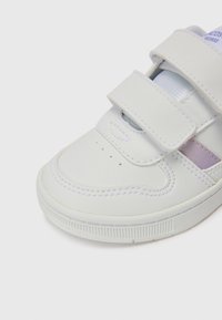 Baskets blanches en simili cuir, avec deux bandes Velcro, des perforations sur le bout et une bande d'accent violet clair.