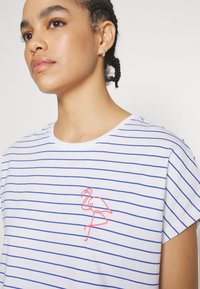 White t-shirt with blue horizontal stripes and short sleeves. Features a pink flamingo embroidery on the left side. Smooth cotton texture.