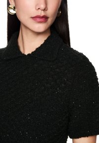 Schwarzes, strukturiertes Strickoberteil mit Kragen und dezenten Verzierung, kurzärmlig. Goldene Statement-Ohrringe sind sichtbar.