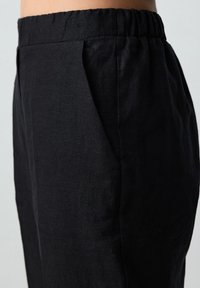 Schwarze Leinenhose mit einem elastischen Bund, seitlichen Falten und einer glatten Textur, die eine taillierte Passform und ein minimales Design betont.