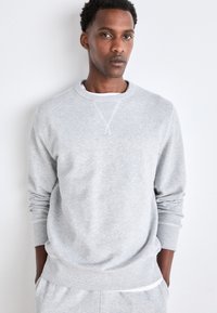 Gray cotton crewneck sweatshirt with long sleeves, ribbed cuffs, and textured fabric. Worn over a white t-shirt, minimal design.