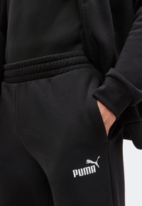 Personne portant un pantalon de survêtement PUMA noir avec la main dans la poche, vue partielle de la veste assortie, logo PUMA blanc visible sur la cuisse gauche.